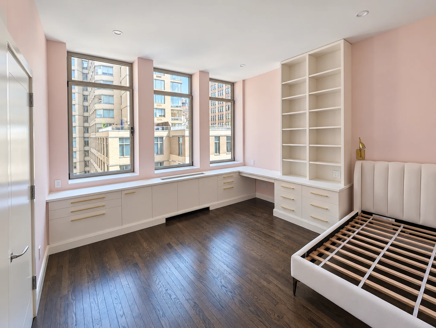 Painted fitted cupboards and drawers beneath three windows, meeting a tall bookcase in a Chelsea room