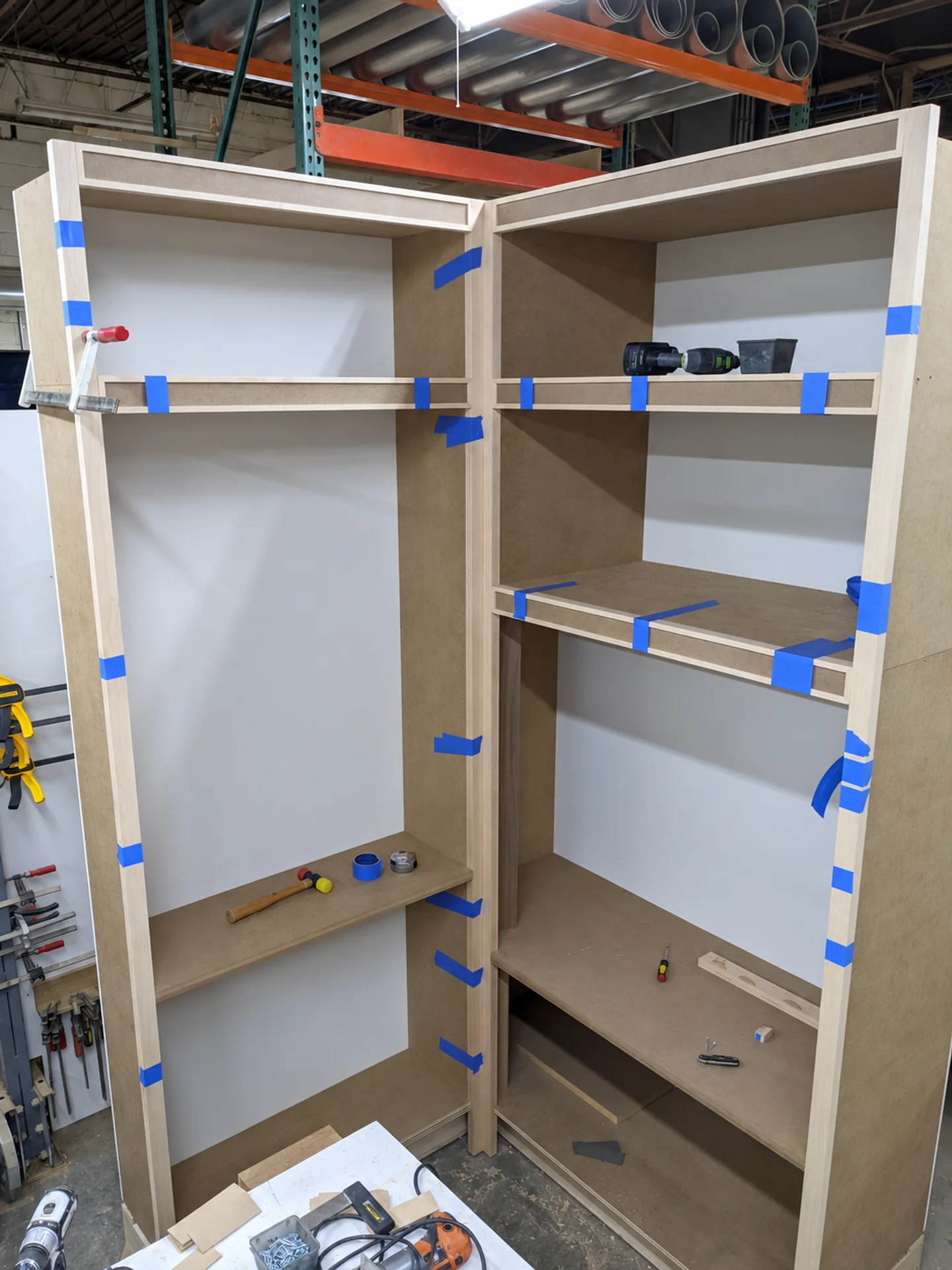 Unfinished corner cabinetry being fitted in the JQ workshop, with clamps, blue assembly tape and a drill on the shelf